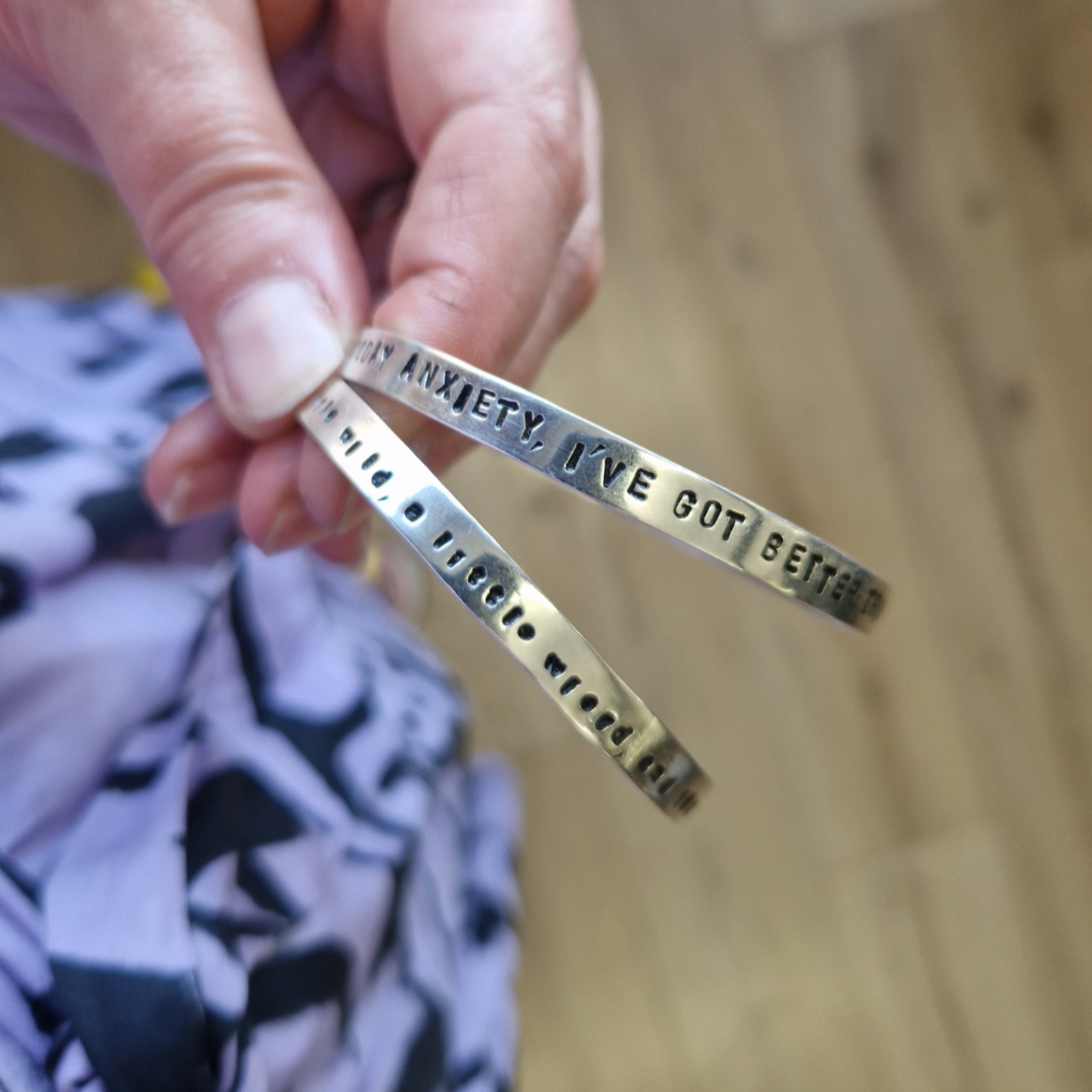 Two silver bracelets with stamped text held by a hand on a wooden surface.