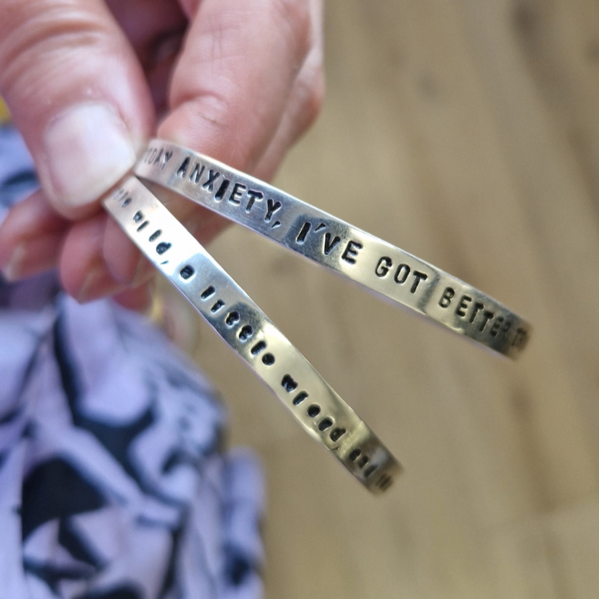Two silver bracelets with stamped text held by a hand on a wooden surface.