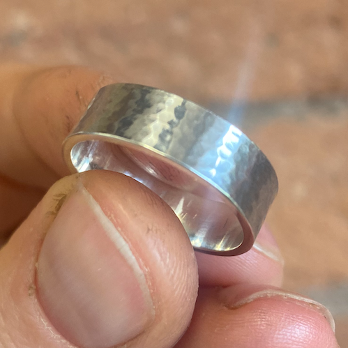 A hand holding a chunky hammered silver ring against a brick wall background.
